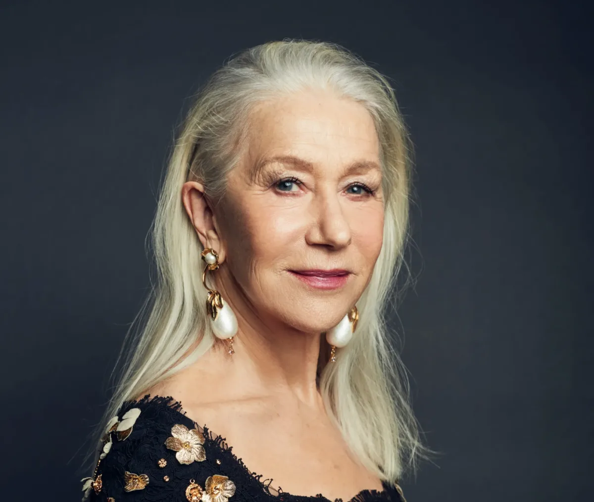 Retrato de busto de la actriz Helen Mirren posando con una sonrisa sutil frente a un fondo oscuro, con motivo de su nombramiento como receptora del Premio Cecil B. DeMille de los Globos de Oro. Luciendo su característico cabello largo y blanco peinado hacia atrás, viste un elegante vestido oscuro con encaje y detalles florales dorados, complementado con unos pendientes de perla de gran tamaño.