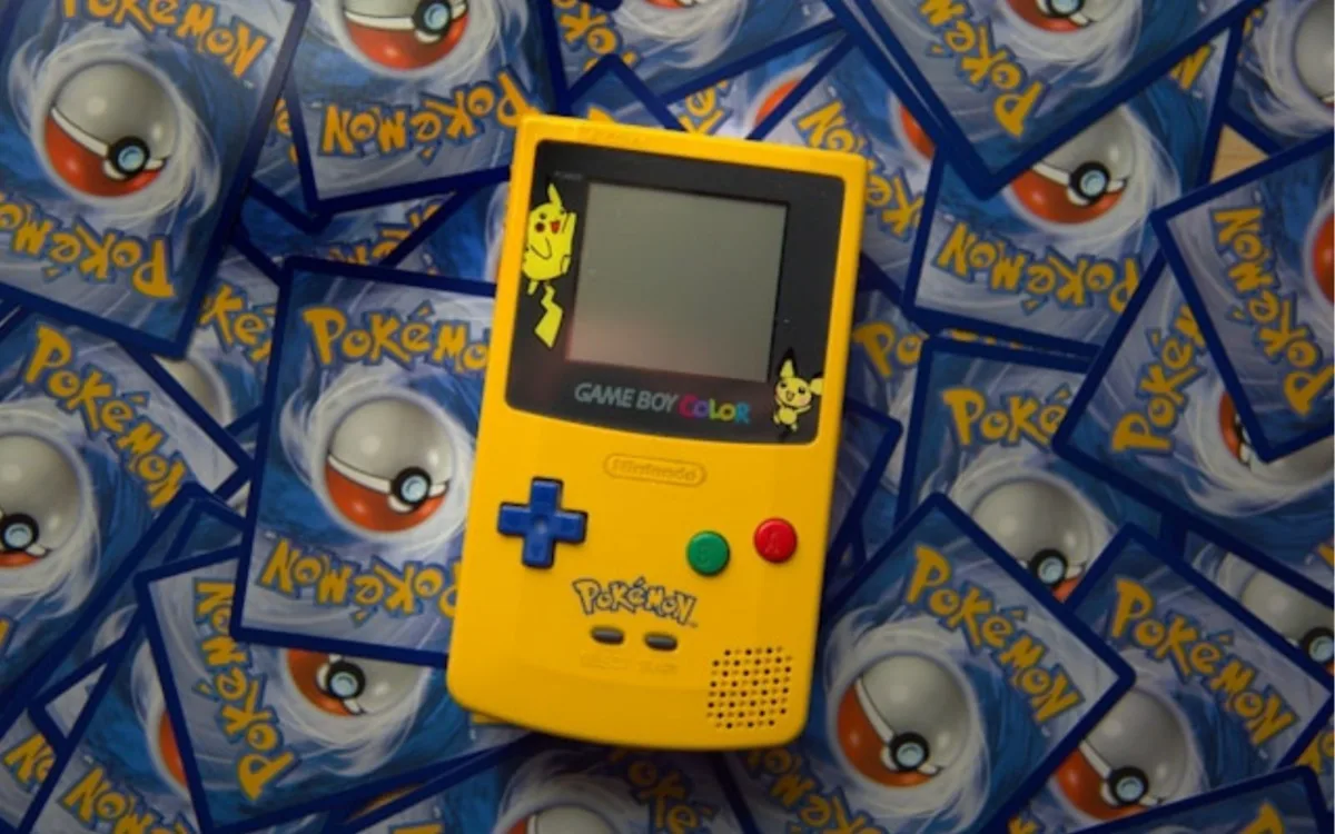 Consola Game Boy Color edición especial de Pokémon en color amarillo, decorada con ilustraciones de Pikachu y Pichu alrededor de la pantalla, colocada sobre un fondo de cartas coleccionables de Pokémon.