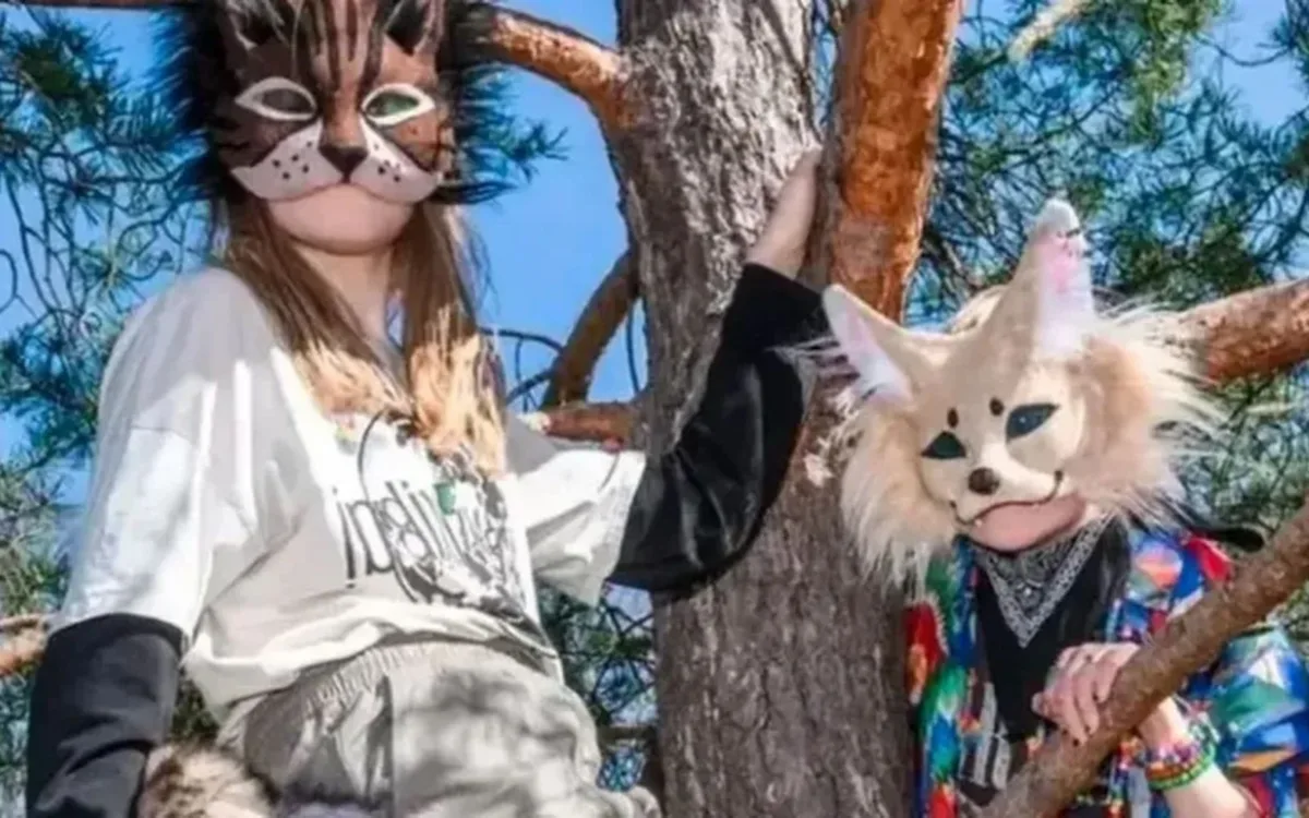 Dos jóvenes therians posan entre las ramas de un pino bajo un cielo azul despejado. Ambos visten máscaras de animales detalladas: a la izquierda, una persona lleva una máscara de felino con orejas puntiagudas y una camiseta blanca; a la derecha, la otra persona luce una máscara de zorro de colores claros y grandes orejas, acompañada de una camisa con un estampado vibrante y múltiples pulseras de colores.