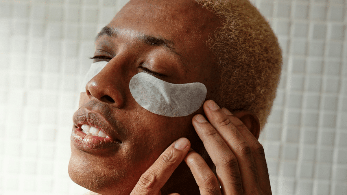 Primer plano de un hombre joven con parches blancos para el contorno de ojos aplicados sobre la piel, mientras cierra los ojos en un momento de relajación.