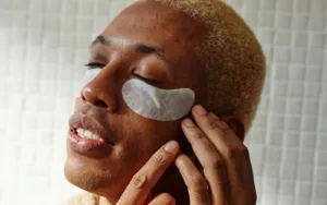 Primer plano de un hombre joven con parches blancos para el contorno de ojos aplicados sobre la piel, mientras cierra los ojos en un momento de relajación.