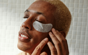 Primer plano de un hombre joven con parches blancos para el contorno de ojos aplicados sobre la piel, mientras cierra los ojos en un momento de relajación.