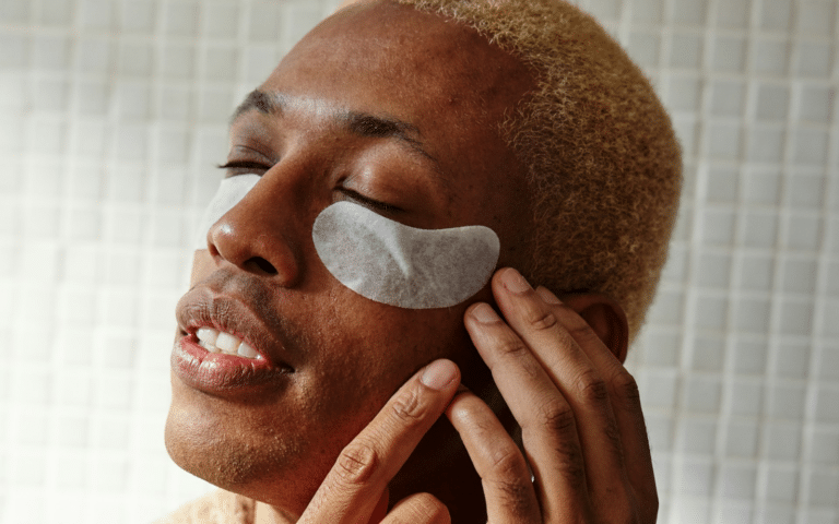 Primer plano de un hombre joven con parches blancos para el contorno de ojos aplicados sobre la piel, mientras cierra los ojos en un momento de relajación.