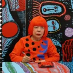 Yayoi Kusama pintando frente a uno de sus característicos fondos llenos de lunares y colores vibrantes